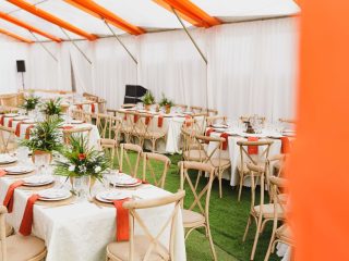 Beautifully arranged event tables with floral centerpieces inside a white and orange tent.
